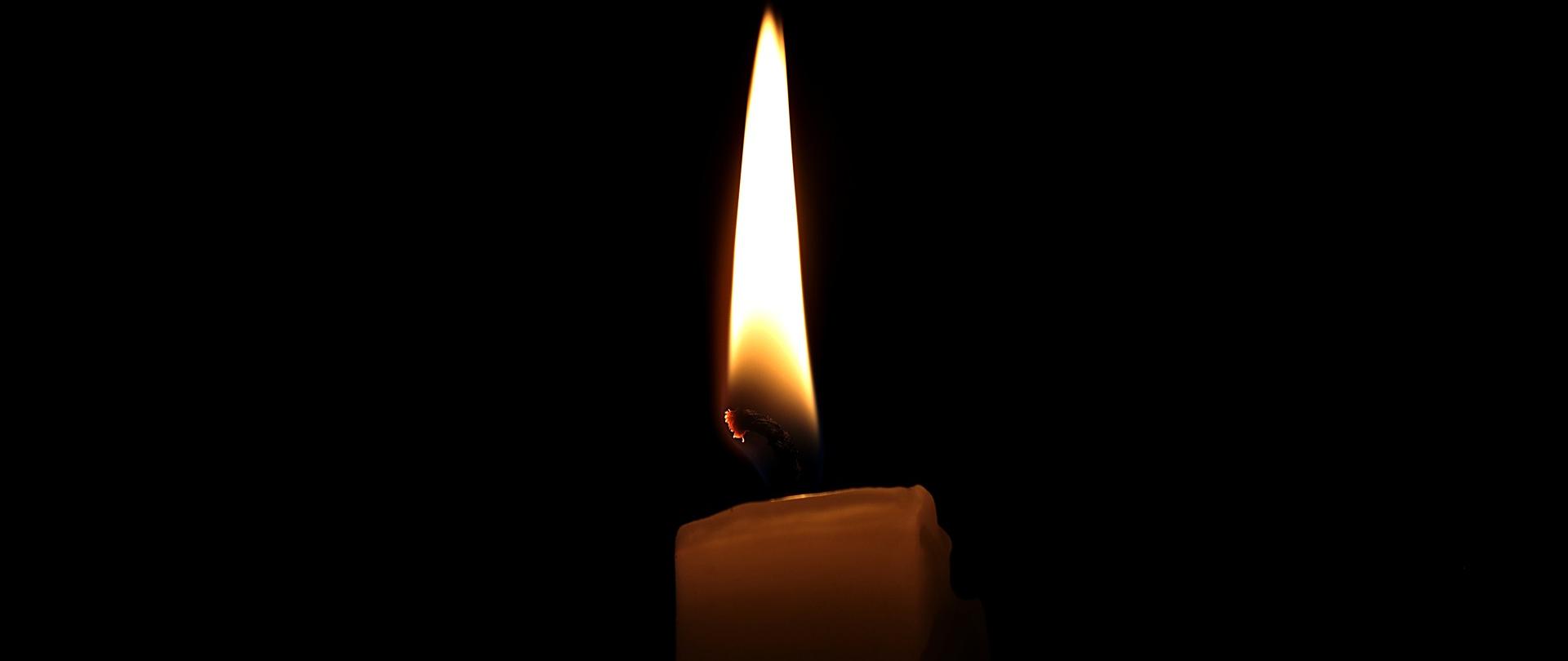 Na zdjęciu w centralnej części kadru świeczka, tlący się płomień. Świeczka jest jasnego koloru ale ogień dodaje lekkiego kolorytu żółci. Tło czarne. 
