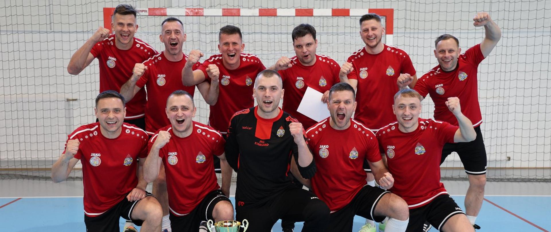 Drużyna sportowa w czerwonych koszulkach pozuje do wspólnego zdjęcia w hali sportowej. Zawodnicy stoją i klęczą przed bramką, unosząc zaciśnięte pięści w geście radości. Na podłodze przed nimi znajdują się puchar, dyplomy oraz piłki.