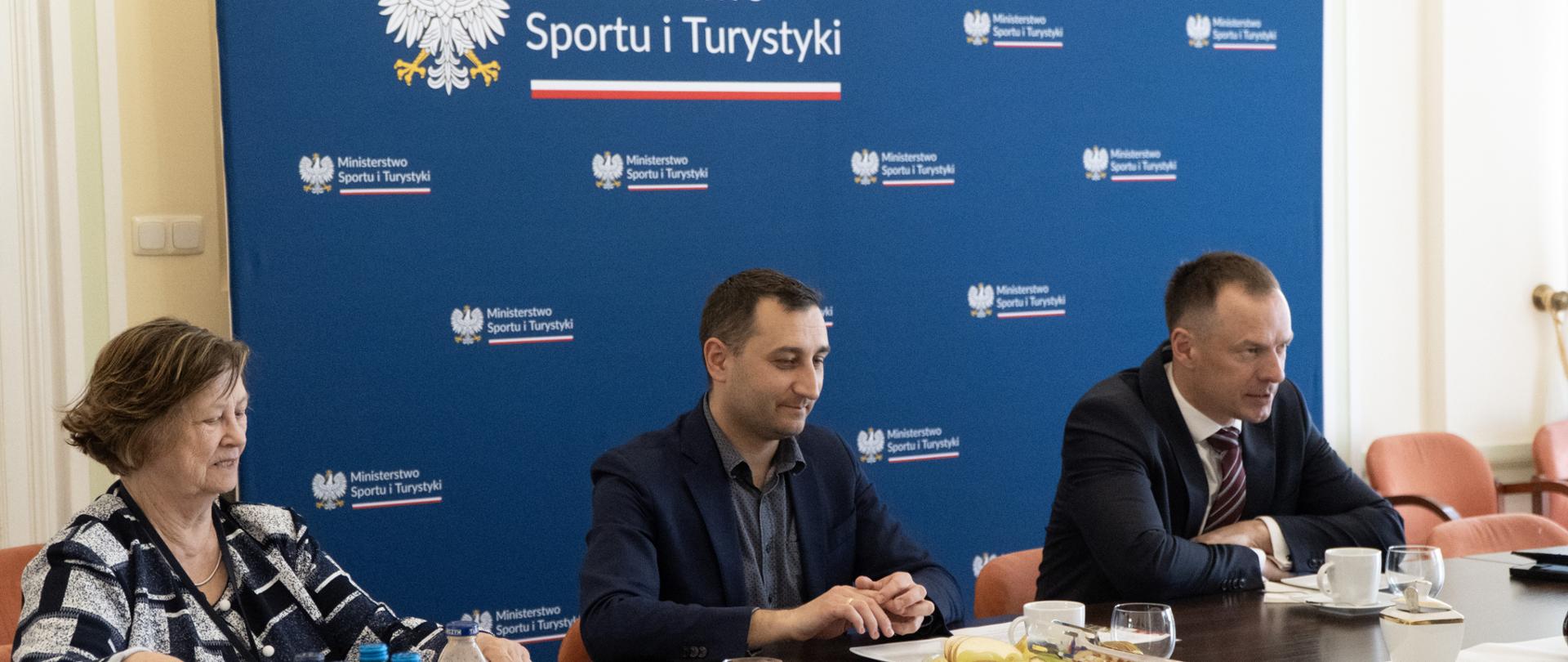 Spotkanie Piotra Borysa ze środowiskiem sektora turystyki campingowej. Na pierwszym planie trzy osoby na tle niebieskiej ścianki z logiem ministerstwa