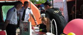 Komendant Powiatowy PSP w Brzezinach oraz Jego Zastępca pod namiotem nadzorują młodzież w rozwiązywaniu testu.