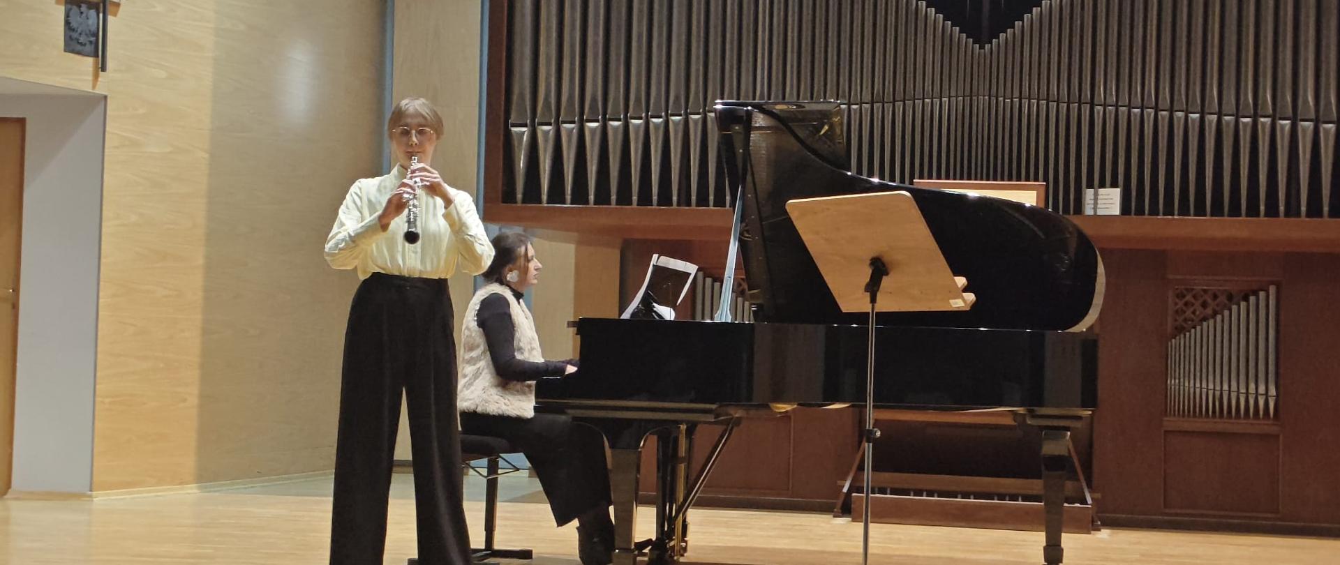 Zdjęcie przedstawia scenę w sali koncertowej podczas występu muzyki kameralnej. W centrum kadru znajdują się dwie młode kobiety i duży, czarny fortepian. Instrumentalistka Dęta (po lewej): Stoi w pozycji frontalnej. Ma jasne, krótkie włosy i okulary. Ubrana jest w jasną, kremową koszulę z długim rękawem oraz ciemne, czarne spodnie z szerokimi nogawkami. Gra na jasnym, podłużnym instrumencie dętym, prawdopodobnie flecie lub klarnecie, który trzyma przy ustach. Jej wyraz twarzy jest skupiony. Pianistka (siedzi przy fortepianie): Siedzi tyłem do lewej strony kadru, za dużym, czarnym fortepianem koncertowym (grand piano). Jest ubrana na ciemno, a na wierzchu ma jasną kamizelkę bez rękawów. Skupia się na partyturze leżącej na pulpicie fortepianu, jej dłonie spoczywają na klawiaturze. Fortepian: Czarny, lśniący, zajmuje znaczną część prawego i środkowego planu. Na fortepianie, zwrócony w stronę publiczności, znajduje się drewniany pulpit na nuty, wsparty na statywie. Tło: Tłem dla występu jest duży prospekt organowy (część organów piszczałkowych) złożony z rzędów drewnianych lub metalowych piszczałek, które nadają scenie uroczysty charakter. Ściany sali koncertowej po bokach są pokryte jasnym drewnem. Scena jest dobrze oświetlona, a światło jest ciepłe, tworząc kontrast między jasnymi ubraniami a ciemnym instrumentem i tłem.