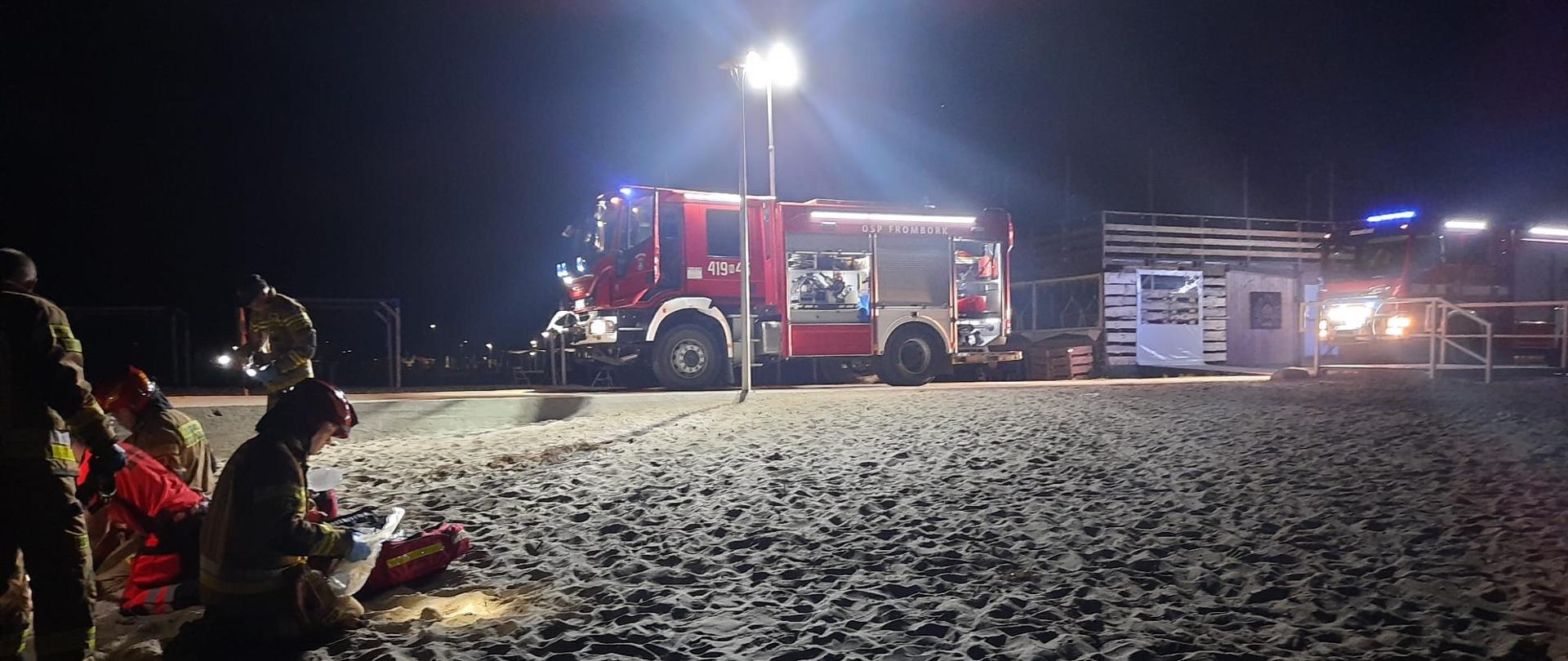 Zdjęcie zrobione nocą. Plaża, teren oświetlany przez samochody strażackie, na pisku grupa strażaków.