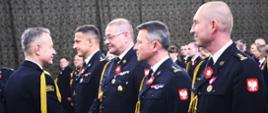 Czterech oficerów PSP stojących w szeregu przed Komendantem Głównym PSP, który gratuluje Zastępcy Komendanta Centralnej Szkoły PSP otrzymanego awansu na stopień starszego brygadiera