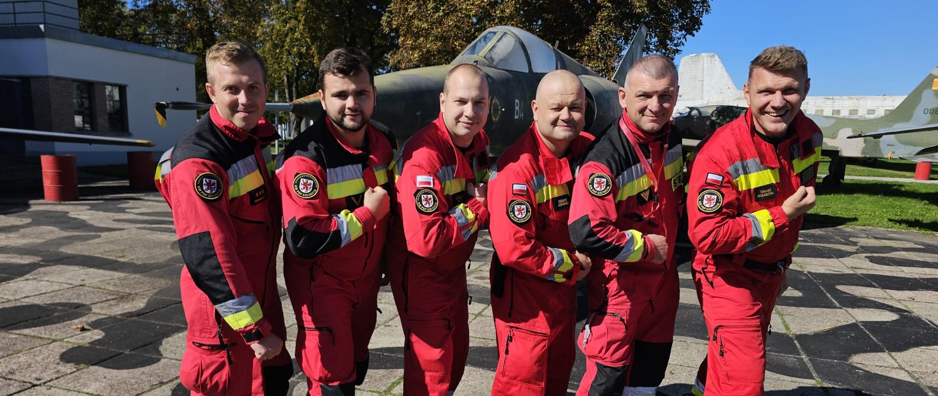 Opis przedstawia sześciu mężczyzn w czerwonych kombinezonach, stojących obok siebie w rzędzie na zewnątrz. Wszyscy są uśmiechnięci i patrzą w kierunku aparatu. Tło zdjęcia stanowią dwa samoloty wojskowe. Na ziemi widać czarne, okrągłe płyty. Mężczyźni mają na sobie czerwone kombinezony z czarnymi i żółtymi wstawkami. Na rękawach widoczne są naszywki z polską flagą. Każdy z mężczyzn ma inny gest, niektórzy zaciskają pięści, inni mają ręce skrzyżowane. W tle, za mężczyznami, znajdują się dwa zielone samoloty wojskowe. Jeden z nich, znajdujący się w centrum, ma pod skrzydłami rakiety. Drugi, widoczny częściowo po prawej stronie zdjęcia, ma na ogonie biało-czerwoną szachownicę. Całość zdjęcia oświetla jasne, słoneczne światło, a niebo w tle jest bezchmurne i niebieskie.