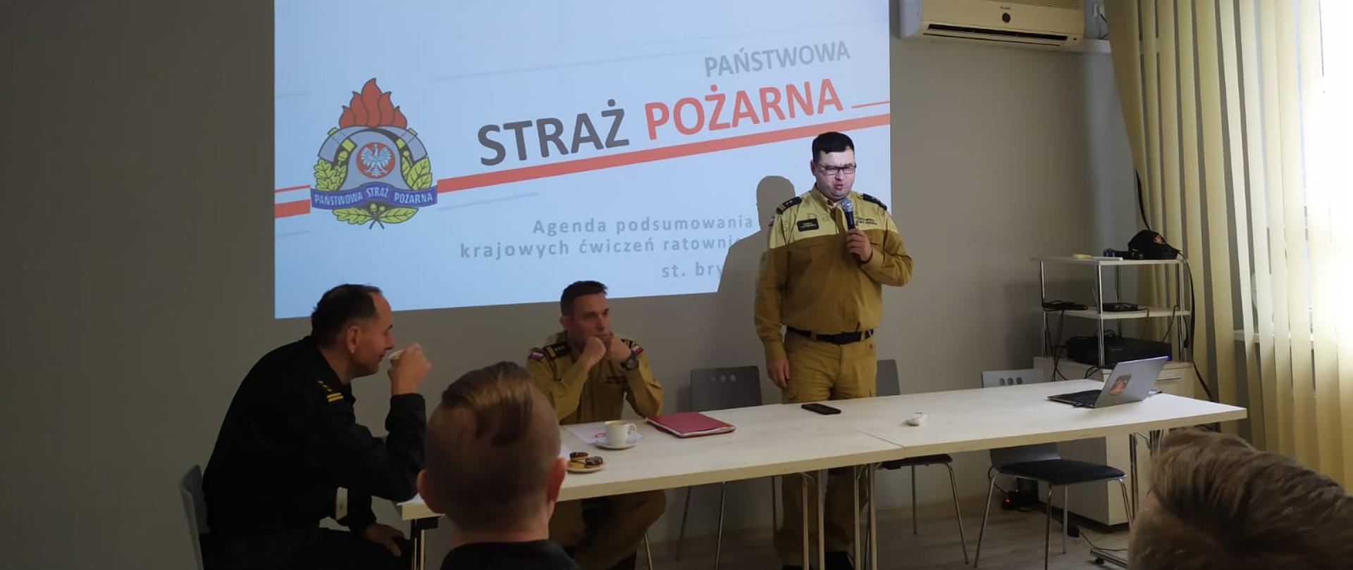 trzech strażaków siedzi przy stole, jeden stoi z mikrofonem, w tle slajd prezentacji z napisem państwowa straż pożarna