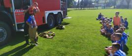 Instruktaż ubierania ubrania specjalnego na trawiastym boisku. Dzieci w półkolu słuchają funkcjonariusza PSP, który prezentuje proces na jednym chłopcu. W tle auto pożarnicze.
