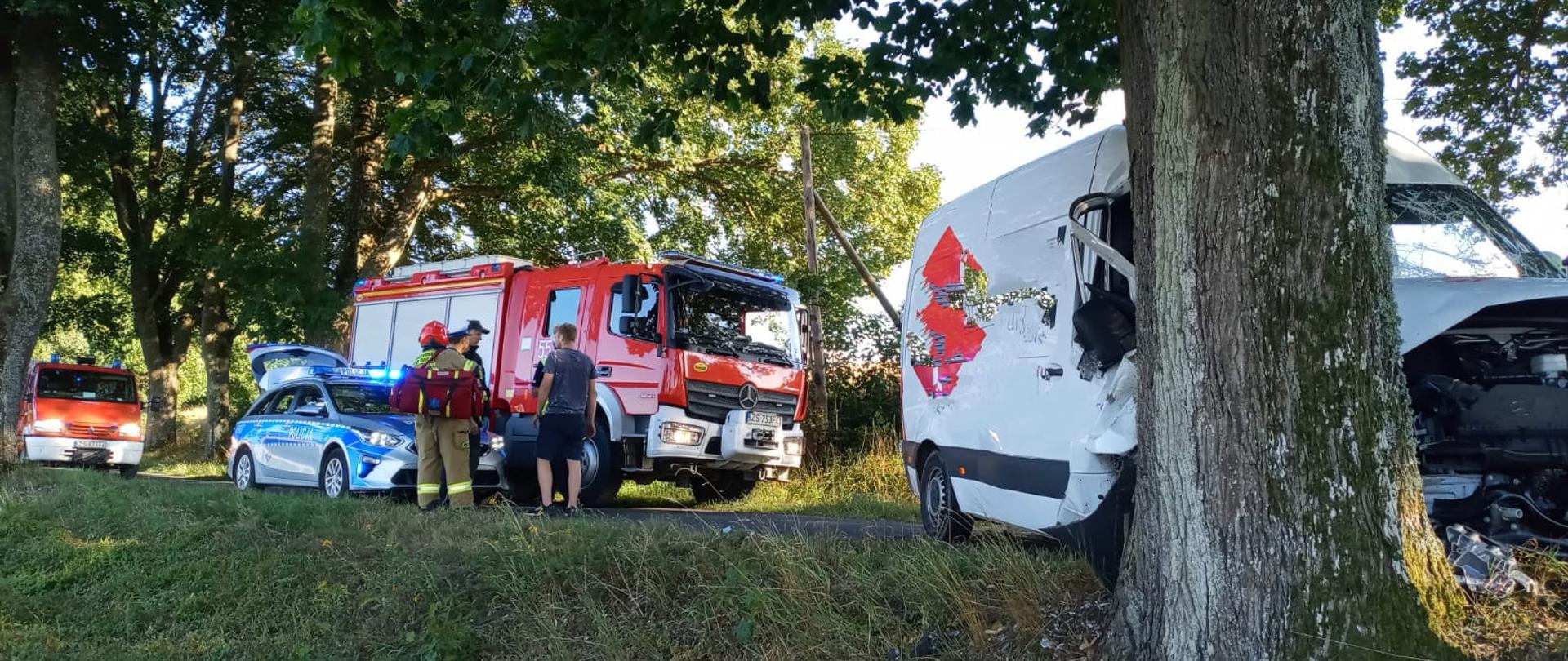 Zdjęcie przedstawia biały samochód dostawczy z rozbitym przodem, który znajduję się przed drzewem. W tle widać samochody służb ratowniczych: straż pożarna oraz policja.