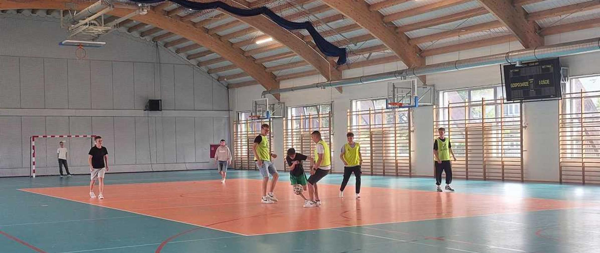 Zdjęcie przedstawia wnętrze nowoczesnej, jasnej hali sportowej, w której grupa nastolatków gra w piłkę nożną.Architektura i wnętrzeDach: Wysoki, łukowy strop z drewnianych konstrukcji i jasnych paneli.Oświetlenie: Pod dachem zamontowano długie, podłużne lampy dające jasne światło.Okna: Duże przeszklenia na bocznej ścianie wpuszczają naturalne światło dzienne.Ściany: Jasnoszare, z zamontowanymi drabinkami gimnastycznymi i koszami do koszykówki.Tablica: Elektroniczna tablica wyników w prawym górnym rogu z napisami „GOSPODARZE” i „GOŚCIE”.Nawierzchnia i liniePodłoga: Sportowa, gumowa lub z tworzywa sztucznego.Strefy: Centralna część boiska ma kolor jasnopomarańczowy, a otaczające ją tło jest jasnoniebieskie.Linie: Wyraźne, namalowane na podłożu linie w kolorze czerwonym, białym i czarnym wyznaczają granice różnych boisk.Ludzie i akcjaGracze: Około ośmiu chłopców w wieku szkolnym, ubranych w stroje sportowe (krótkie spodenki, koszulki, buty sportowe).Znaczniki: Część zawodników ma na sobie jaskrawe, żółto-zielone kamizelki treningowe.Akcja: W centralnej części pomarańczowego pola skupiona jest grupa czterech chłopców walczących o piłkę. Jeden z nich próbuje ją dryblować nogą. Pozostali gracze są rozproszeni po boisku, obserwując akcję.Tło: Po lewej stronie w głębi widać biało-czerwoną bramkę do piłki ręcznej/halowej.