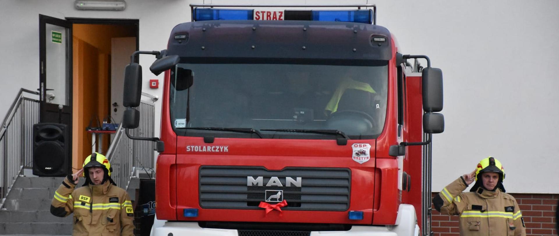 Po prawej i lewej stronie wozu strażackiego stoją na baczność druhowie OSP. Do wozu przyczepiona jest czerwona wstążka.
