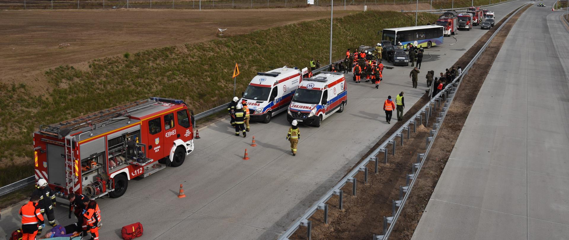 Pozorowany wypadek autokaru i samochodów osobowych. Wokół pojazdy pogotowia i straży pożarnej. Strażacy i ratownicy medyczni ćwiczą udzielanie pomocy poszkodowanym.