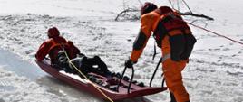 
Na zdjęciu widoczni dwaj strażacy na skutym lodzie akwenie wodnym. Obaj w suchych skafandrach koloru pomarańczowego w czerwonych kaskach ochronnych. Jeden ratownik leży na saniach drugi pchający sanie oparty o podpory udają się do osoby poszkodowanej w celu ewakuacji osoby poszkodowanej z wody.