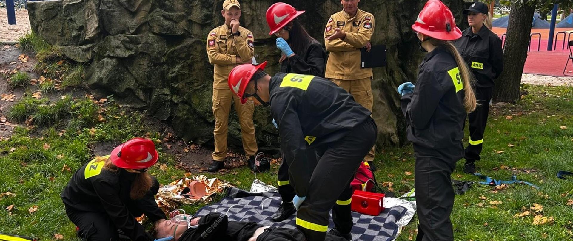 Na zdjęciu Młodzieżowa drużyna Pożarnicza podczas wykonywania jednego z zadań obok funkcjonariusze PSP który oceniacy zadanie, oraz osoba poszkodowana na kocu