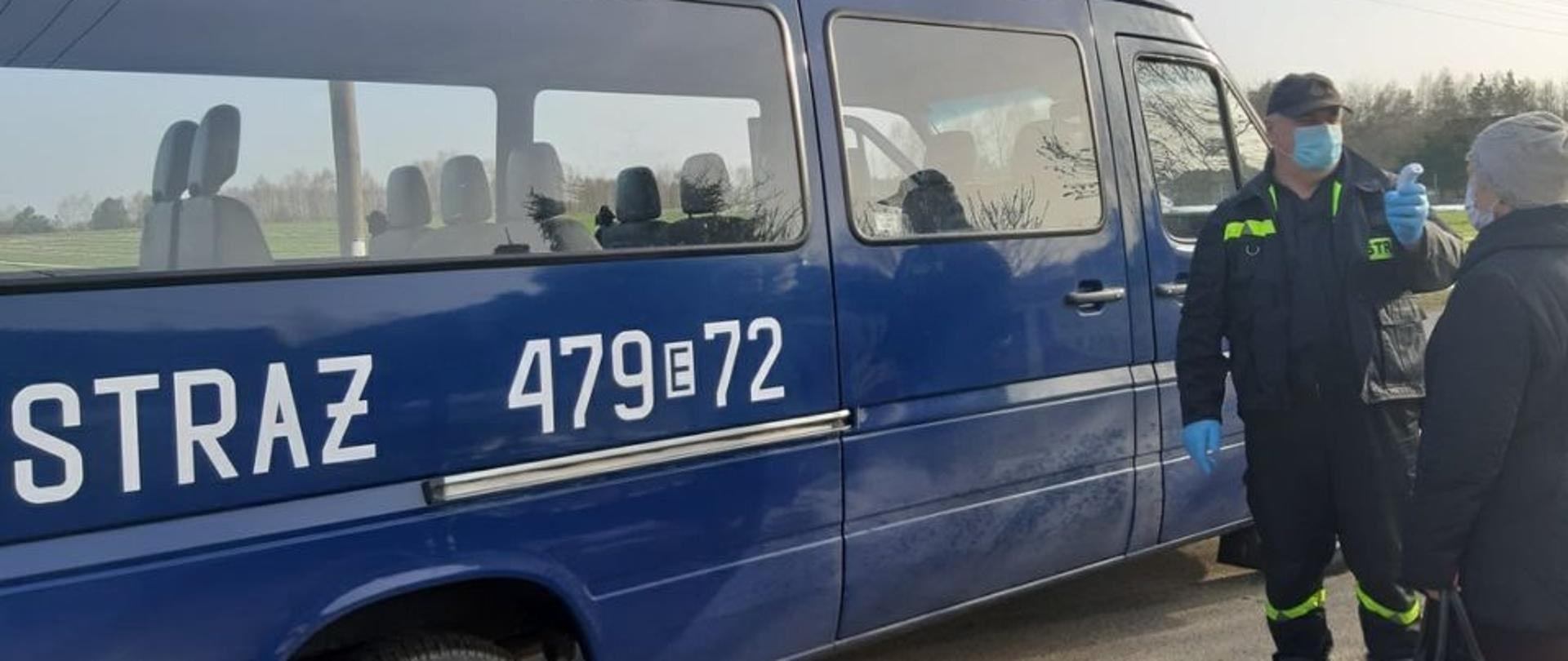 Zdjęcie przedstawia strażaka OSP w ubraniu koszarowym wykonującego pomiar temperatury osobie starszej. W tle pojazd pożarniczy typu mikrobus. Wszyscy mają założone maseczki.