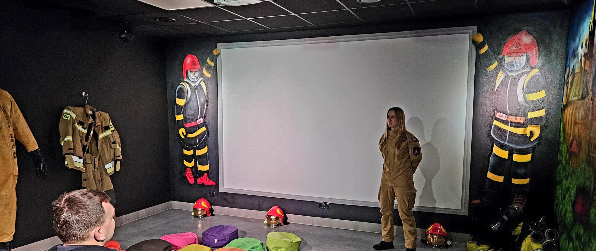 Zdjęcie pokazuje strażaczkę podczas prezentacji. Na podłodze leżą kolorowe poduszki do siedzenia , na ścianach są rysunki strażaków. 