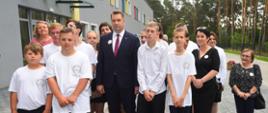 Na dworze przed szarym budynkiem stoi minister Czarnek w otoczeniu kilkunastu młodych ludzi w białych koszulkach.