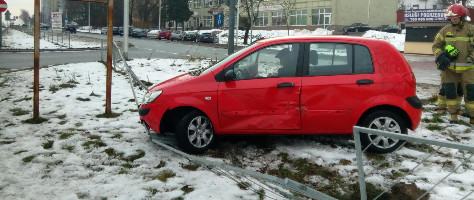 Na pierwszym planie widać czerwony samochód z uszkodzonym bokiem, który stoi na zniszczonych barierkach, a za nim znajduje się strażak.