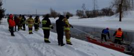 Dwóch strażaków ubranych w kapoki na łodzi prowadzących działania poszukiwawcze na rzece Nurzec. Na brzegu pozostali strażacy biorący udział w akcji.