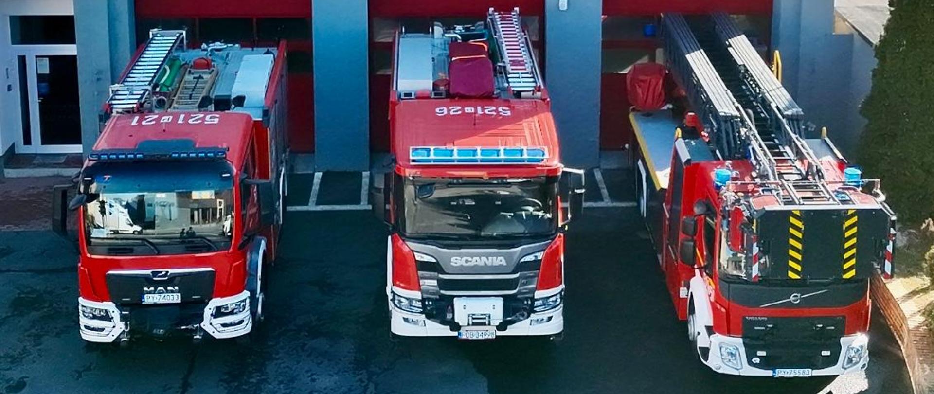 Trzy czerwone wozy strażackie zaparkowane w garażach remizy strażackiej. Pojazdy stoją obok siebie w otwartych bramach budynku z szarą elewacją i czerwonymi drzwiami garażowymi. Na jednym z wozów widoczna jest drabina mechaniczna