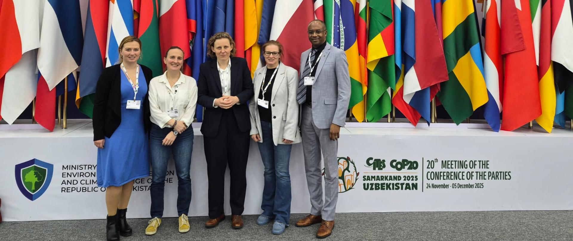 Grupa 5 osób (4 kobiety i 1mężczyzna) w oficjalnych strojach stoi na ściany utworzonej z flag różnych państw na drzewcach - po prawej napis informujący o tym, że to 20. Konferencja na rzecz Dzikiej Przyrody, trwająca od 24 listopada do 5 grudnia 2025 w Samarkandzie w Uzbekistanie 