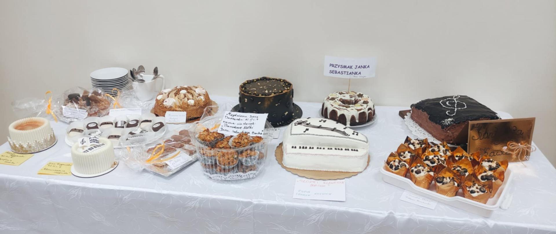 Na zdjęciu widać długi stół przykryty białym obrusem zastawiony różnego rodzaju domowymi wypiekami i słodkościami. Niektóre z nich są z polewą czekoladową czy lukrem, jedno z nich ozdobione jest motywem klucza wiolinowego, inne ma symbol pięciolinii i nut. Na stole stoją też filiżanki i talerzyki