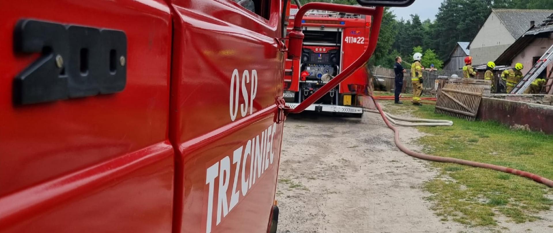 Zdjęcie na pierwszym planie przedstawia samochód rat6owniczo-gasniczy na którym widnieje napis OSP Trzciniec. Na drugim palnie widać budynek inwentarski po pożarze dachu. Strażacy w umundurowaniu specjalnym pracują przy usuwaniu skutków pożaru. 
