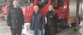 2 strażaków w ubraniach koszarowych stoją wraz ze starszym mężczyzną w ubraniu cywilnym w tle samochody pożarnicze.