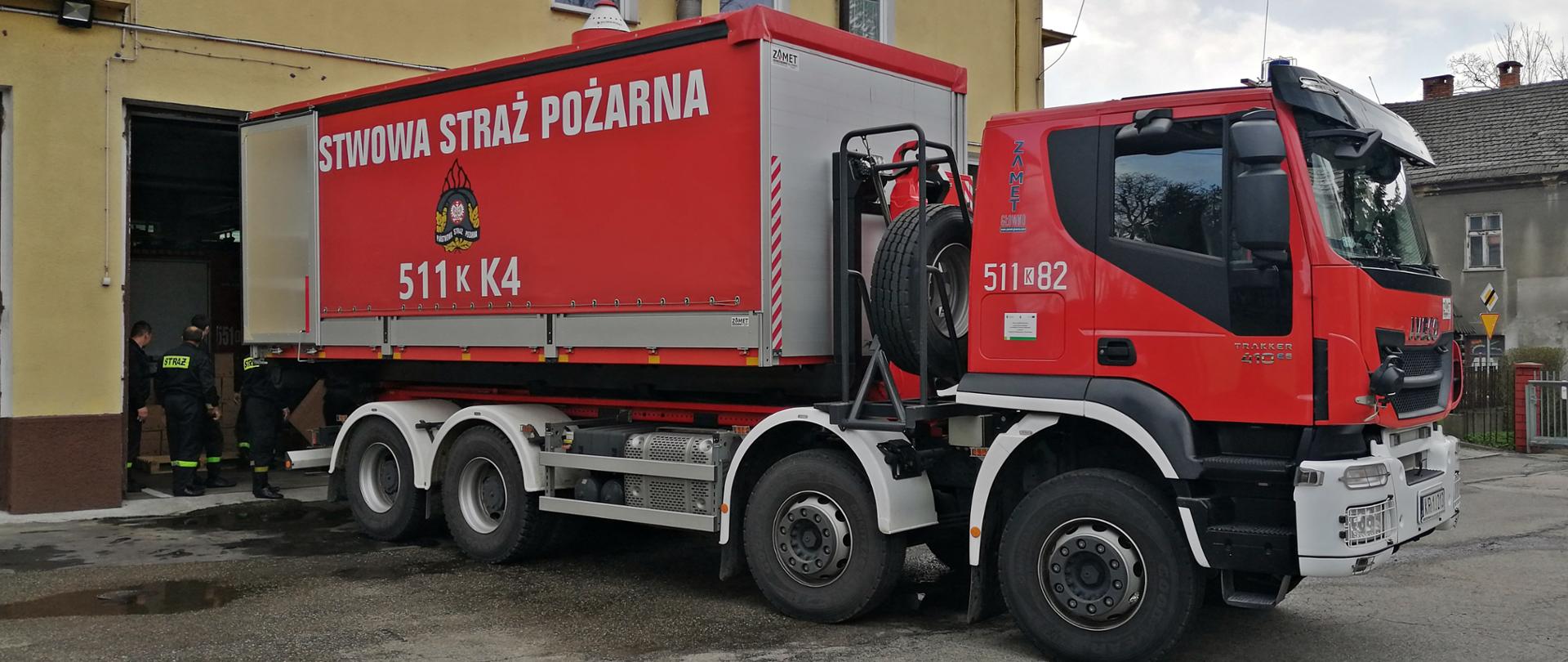 Samochód ciężarowy KP PSP w Oświęcimiu w kolorze czerwonym stoi tyłem do garażu Jednostki Ratowniczo-Gaśniczej w Wadowicach w trakcie rozładunku płynów dezynfekcyjnych. 