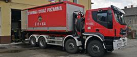 Samochód ciężarowy KP PSP w Oświęcimiu w kolorze czerwonym stoi tyłem do garażu Jednostki Ratowniczo-Gaśniczej w Wadowicach w trakcie rozładunku płynów dezynfekcyjnych. 