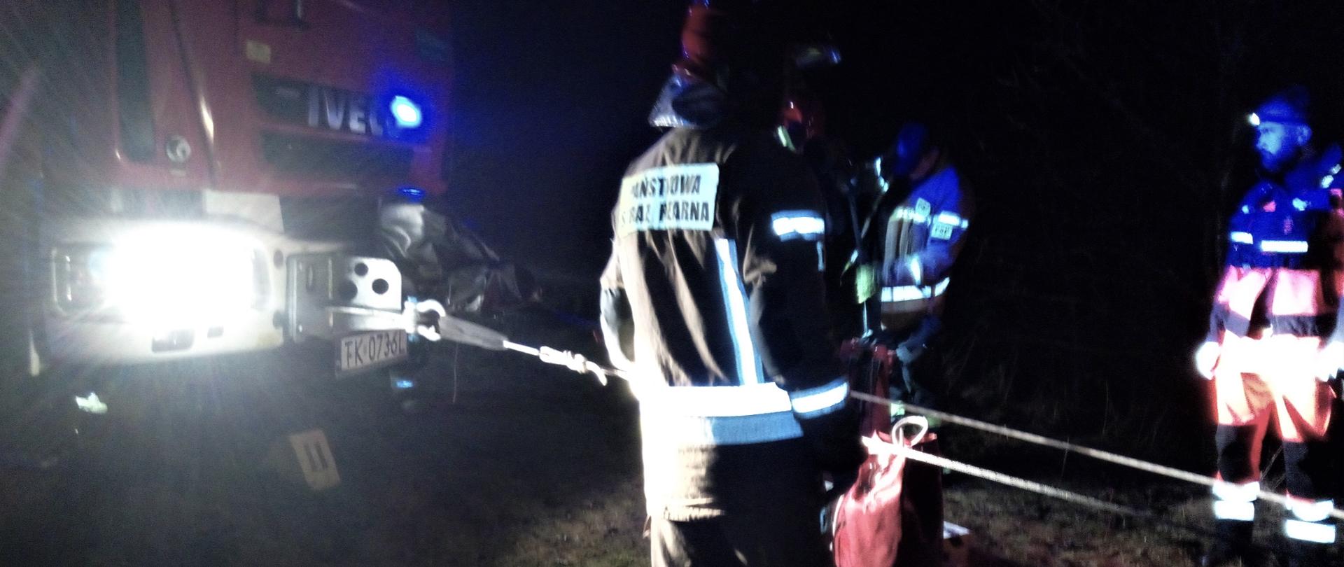 Na zdjęciu przedstawiono przód samochodu pożarniczego, na bazie którego zbudowano stanowisko zjazdowe służące do wyciągnięcia osoby poszkodowanej z dna kamieniołomu. Widzimy trzech ratowników asekurujących stanowisko.