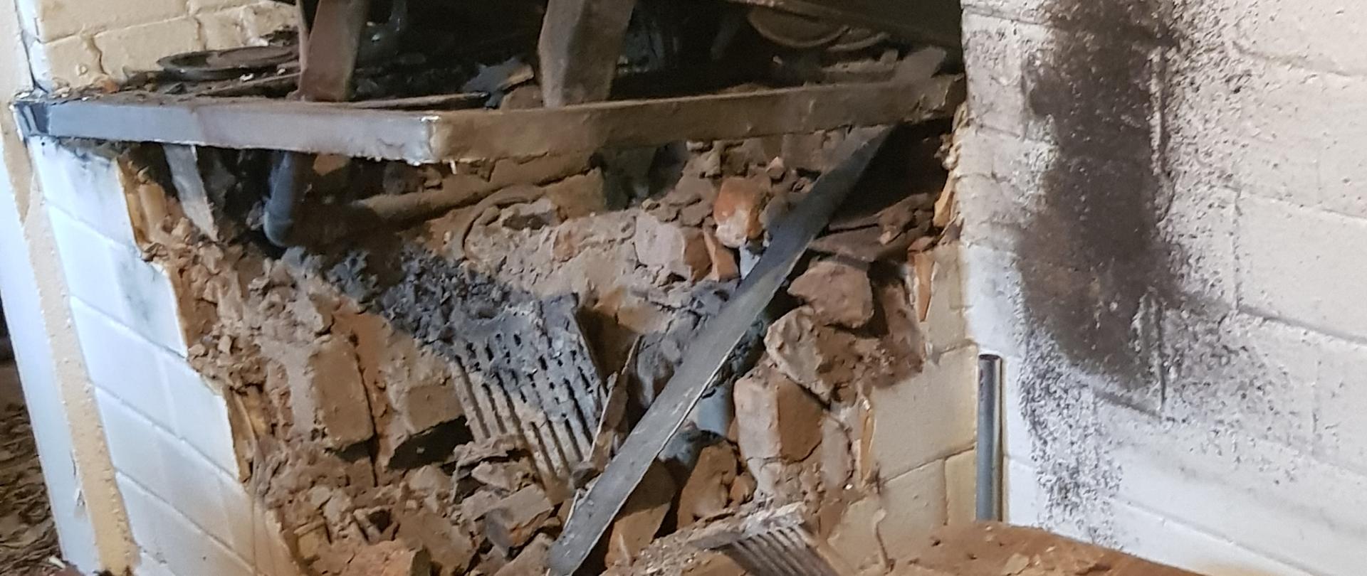 Zniszczony piec kaflowy na skutek rozsadzenia wężownicy. Widoczne liczne kawałki elementów pieca na ziemi oraz wyrwane palenisko.