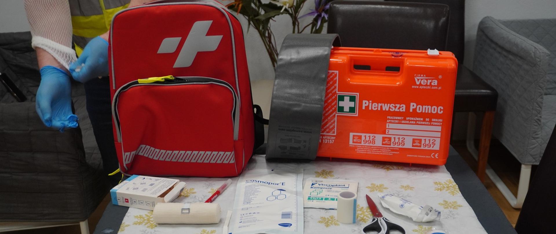 Na stoliku znajduję się plecak, apteczka pierwszej pomocy, szyna, nożyczki, plaster, kompresy, opatrunki oraz bandaż elastyczny.