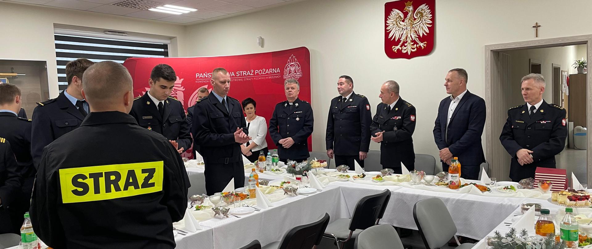 Sala konferencyjna w siedzibie komendy państwowej straży pożarnej w brzezinach zastawione stoły jedzeniem wigilijnym, przy stołach stoją strażacy śpiewają kolędy .