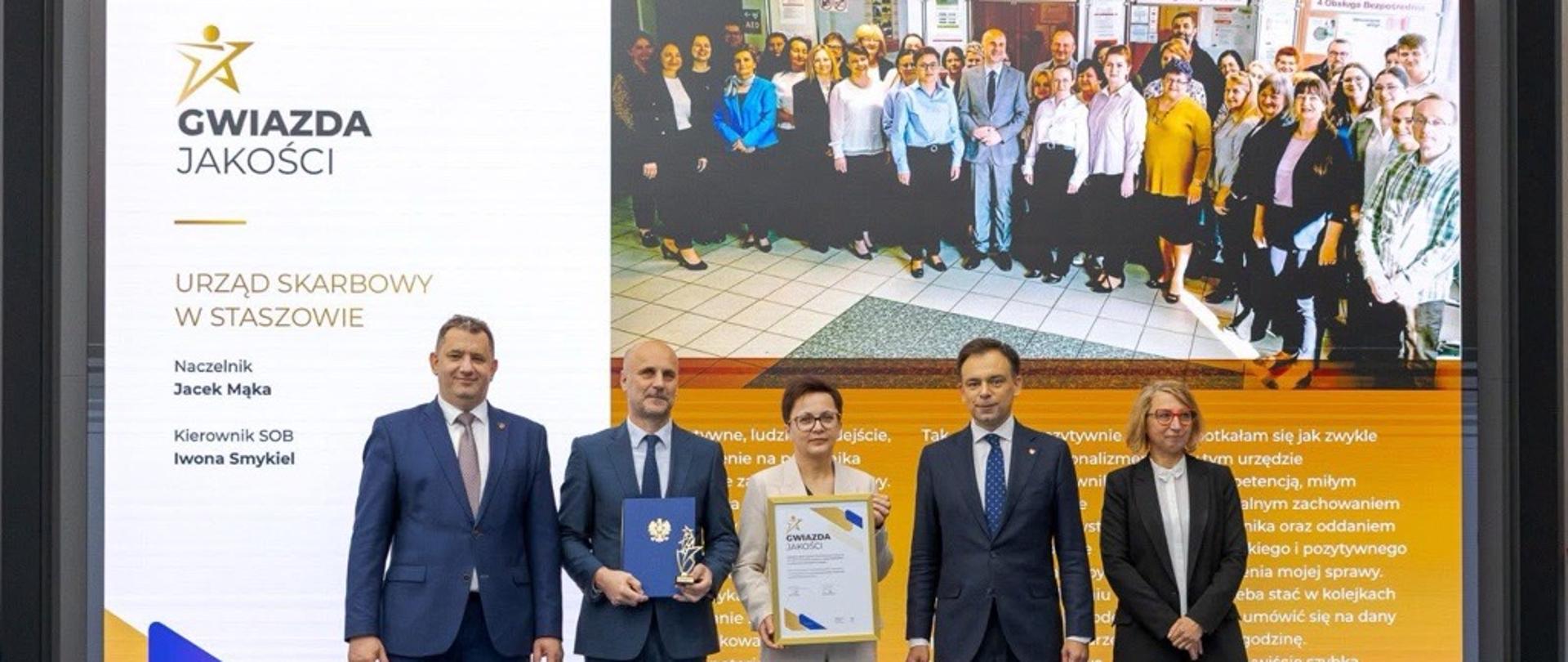 Andrzej Domański, minister finansów i gospodarki, Marcin Łoboda, szef Krajowej Administracji Skarbowej, oraz przedstawiciele Urzędu Skarbowego w Staszowie prezentują dyplom i statuetkę „Gwiazda Jakości”. Na ekranie w tle widoczna jest nazwa urzędu, zdjęcie zespołu oraz fragmenty opinii klientów.