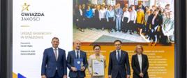 Andrzej Domański, minister finansów i gospodarki, Marcin Łoboda, szef Krajowej Administracji Skarbowej, oraz przedstawiciele Urzędu Skarbowego w Staszowie prezentują dyplom i statuetkę „Gwiazda Jakości”. Na ekranie w tle widoczna jest nazwa urzędu, zdjęcie zespołu oraz fragmenty opinii klientów.