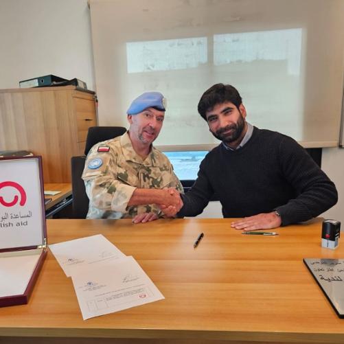 Dwóch mężczyzn ściska sobie rękę przy biurku pozując do zdjęcia, jeden ma błękitny beret i mundur w polskim kamuflażu z polską flagą, przed nimi pudełko na dokumenty z logo Polska pomoc i podpisana umowa