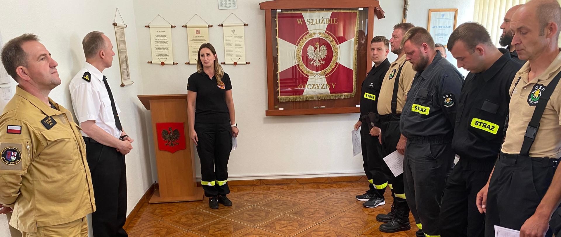 Zdjęcie przedstawia Zakończenie szkolenia kierującego działaniem ratowniczym dla strażaka ratownika Ochotniczych Straży Pożarnych (Dowódcy OSP). strażacy stoją w dwu szeregu ze świadectwami w ręku na środku kobieta przemawia.