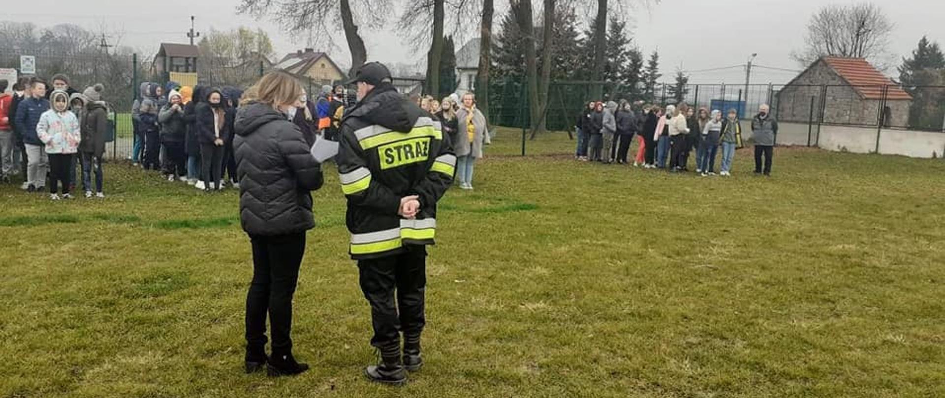 Teren zieleni przed budynkiem szkoły. Stojący tyłem strażak w umundurowaniu koszarowym i w kurtce z elementami odblaskowymi, rozmawia ze stojącą po jego lewej stronie kobietą. Na przeciwko strażaka i kobiety stoi grupa dzieci ustawionych w rzędach.