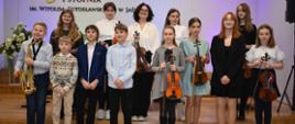 Grupa uczniów w strojach galowych trzyma w rękach swoje instrumenty oraz pozuje do zdjęć. W tle – logo szkoły oraz Godło. Przeważająca kolorystyka – czerń oraz barwy pastelowe.