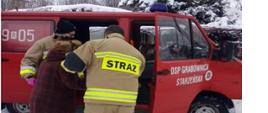 Kolorowe zdjęcie przedstawia na pierwszym planie, czerwony, oznakowany samochód lekki Ochotniczej Straży Pożarnej z napisem OSP Grabownica Starzeńska na otwartych drzwiach. Przy samochodzie, na środku widać 2 umundurowanych strażaków z napisem na plecach Straż, którzy pomagają starszej osobie pośrodku, wejść do samochodu. Na zdjęciu widoczna jest duża ilość śniegu a w tle również widoczne zaśnieżone drzewa.