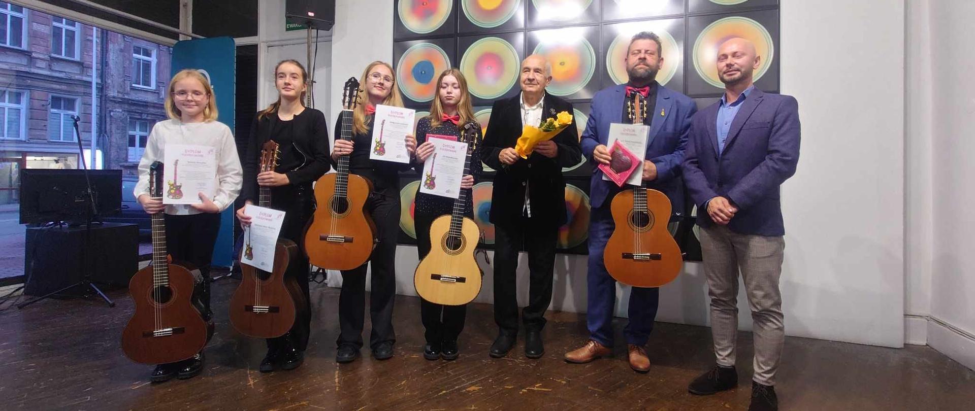Czwórka uczennic trzymająca gitary z dyplomami wraz z trójką osób dorosłych pozujący do zdjęcia grupowego.