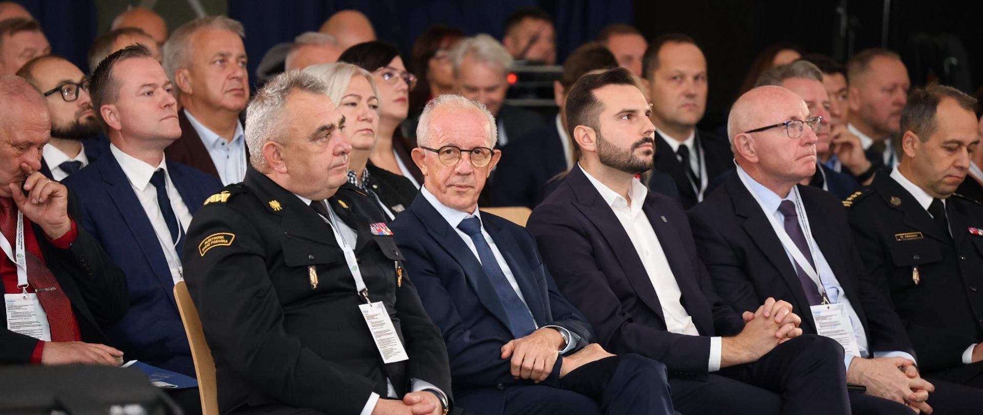 Zdjęcie przedstawia pierwsze rzędy publiczności siedzącej na widowni podczas konferencji. Ludzie siedzą obok siebie, skierowani ku scenie, która znajduje się poza kadrem, w prawym górnym rogu. Wśród publiczności dominują ciemne ubrania: garnitury i mundury, co nadaje wydarzeniu formalny charakter. Tło jest bardzo ciemne, sala jest zaciemniona, a światło skupia się na scenie.