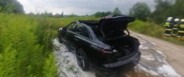 Samochód osobowy stoi na polnej drodze. Na ziemi leży piana gaśnicza. W oddali widać strażaków.