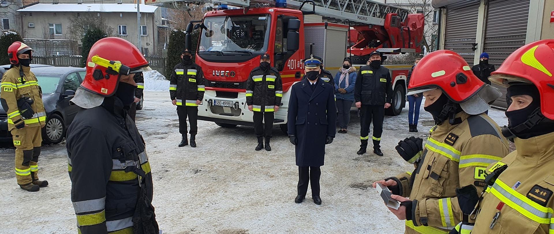 Zdjęcie wykonane na placu Komendy Powiatowej Państwowej Straży Pożarnej na tle samochodu pożarniczego. Komendant Powiatowy mł. bryg. Arkadiusz Spera oraz strażacy Jednostki Ratowniczo - Gaśniczej ubrani w ubranie specjalne żegnają odchodzącego na emeryturę kpt. Janusza Radkowskiego