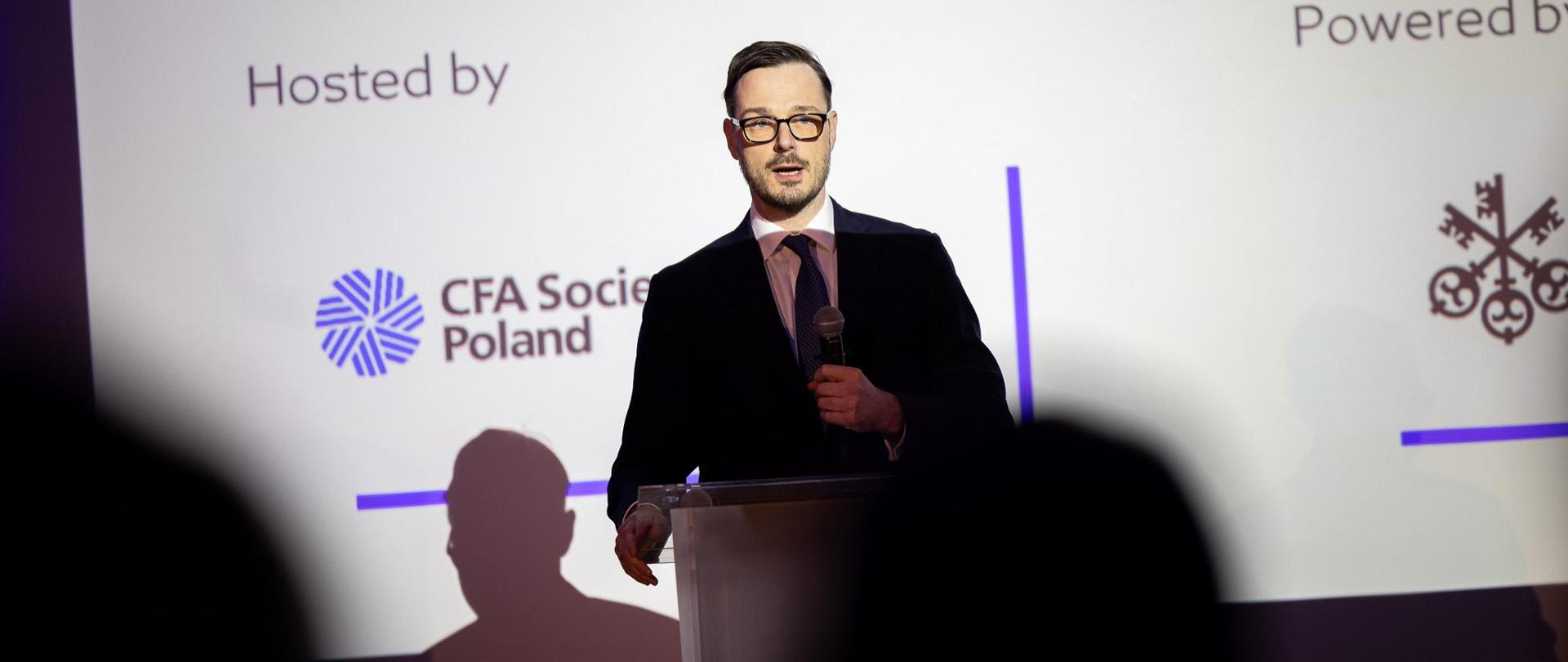 Minister stoi przy mównicy trzymając mikrofon. W tle ekran z nazwą wydarzenia.