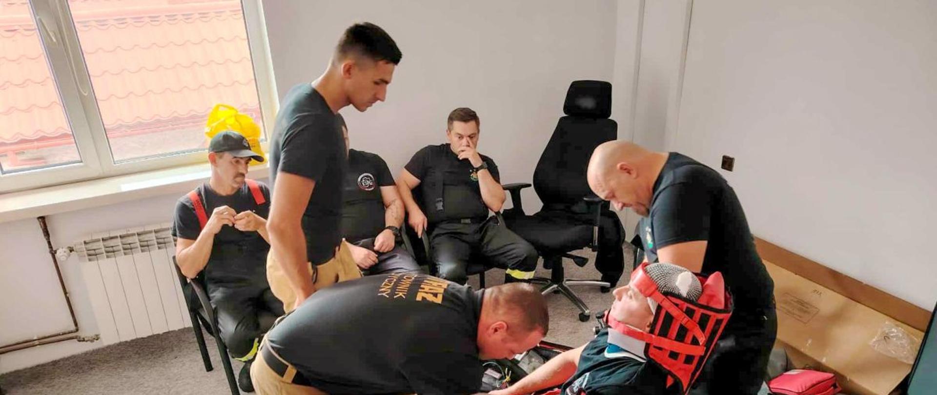 Zdjęcie przedstawia strażaków OSP i PSP. Trzech strażaków zabezpiecza jednego który ubrany ma stabilizator KED. Jeden strażak podchodzi do nich. Widać również torbę r1. Trzech pozostałych strażaków siedzi pod oknem na krzesłach. 