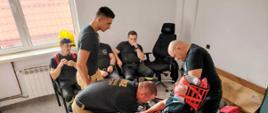 Zdjęcie przedstawia strażaków OSP i PSP. Trzech strażaków zabezpiecza jednego który ubrany ma stabilizator KED. Jeden strażak podchodzi do nich. Widać również torbę r1. Trzech pozostałych strażaków siedzi pod oknem na krzesłach. 
