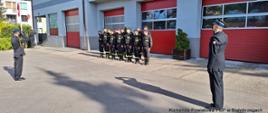 Pododział 16 strażaków w umundurowaniach koszarowych. Zastępca komendanta i dowódca jednostki ratowniczo-gaśniczej oddają honor, fladze Rzeczypospolitej Polskiej, poprzez salutowanie. Po lewej poczet flagowy w trakcie podnoszenia flagi na maszt. W tle budynek Komendy Powiatowej Państwowej Straży Pożarnej w Białobrzegach (szara elewacja, czerwone bramy wyjazdowe)