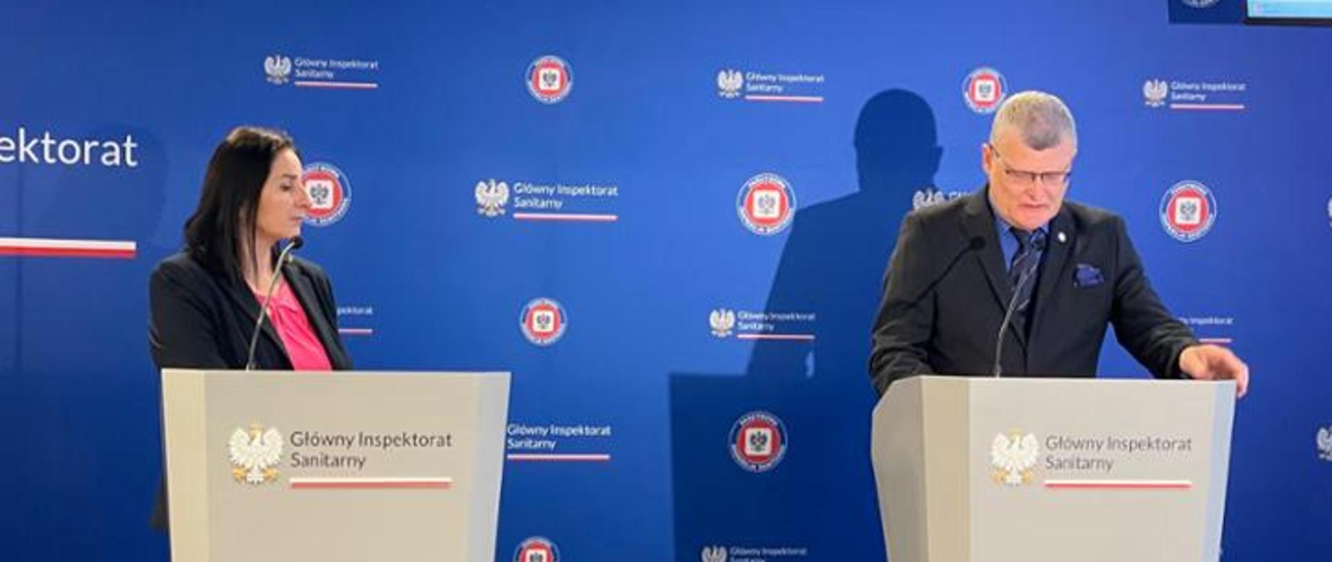 Na zdjęciu widać dwie osoby stojące przy mównicach z logo Głównego Inspektoratu Sanitarnego. Po lewej stronie znajduje się kobieta w ciemnym żakiecie i różowej bluzce, patrząca w stronę mężczyzny po prawej stronie. Tło stanowi niebieska ścianka z oficjalnymi logotypami GIS. Zdjęcie wykonano podczas oficjalnego wystąpienia lub konferencji prasowej.