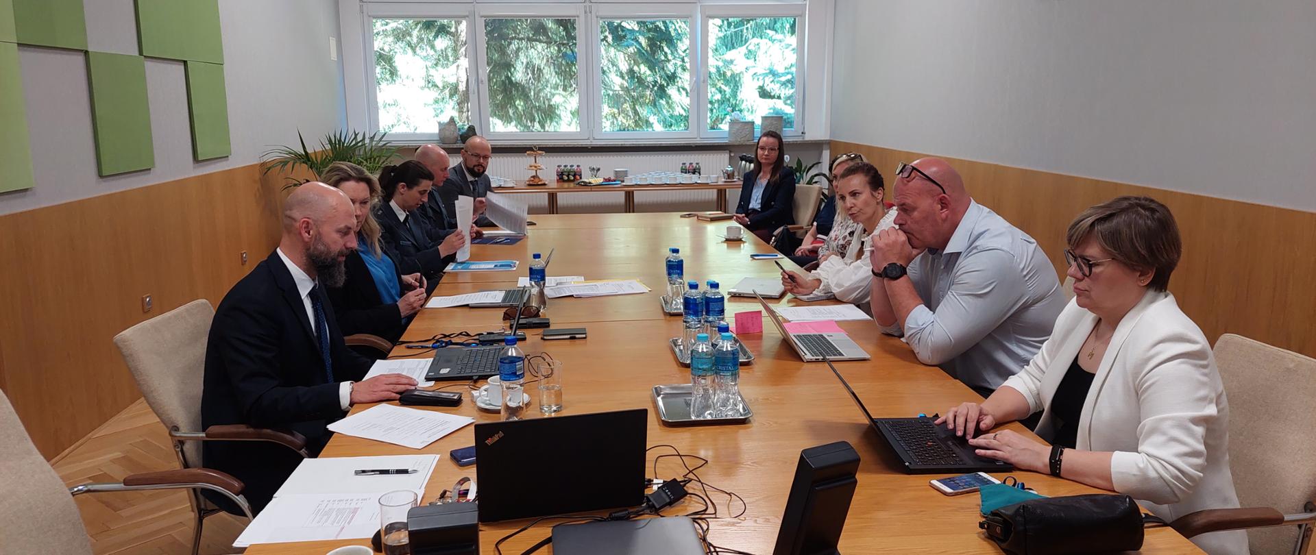 Grupa osób uczestnicząca w spotkaniu. Na pierwszym planie widoczny sprzęt komputerowy. W oddali widać okno. 