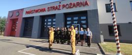 Strażacy stoją w dwuszeregu na tle bram garażowych, po prawej stronie maszt flagowy, przed nim na środku stoi dwóch strażaków w żółtych mundurach - salutują. 
